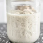 Homemade gluten-free sourdough starter in a glass jar with ingredients.